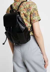 Černý kožený batoh s překlápěcí klopou, stahovací šňůrkou a přední kapsou. Nošený na světlém, dobře padnoucím oblečení.