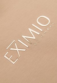 Nom de la marque EXIMIO BY OMBRE embossé en lettres brillantes sur un fond en tissu beige texturé.