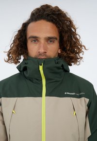 Zeleno-bežová nepremokavá bunda s vysokým golierom, žltým zipsom a zipsovým vreckom. Obsahuje nastaviteľnú šnúrku na kapucni.