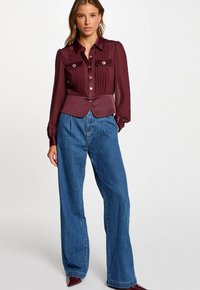Blusa bordeaux a maniche lunghe con pieghe e dettagli con bottoni, abbinata a jeans blu a vita alta e con gamba larga. Tessuto liscio con una vestibilità su misura.