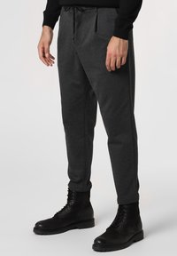 Tmavě šedé jogger kalhoty vyrobené z texturované tkaniny, s elastickým pasem, tkaničkou a postranními švy. V kombinaci s černými šněrovacími botami.