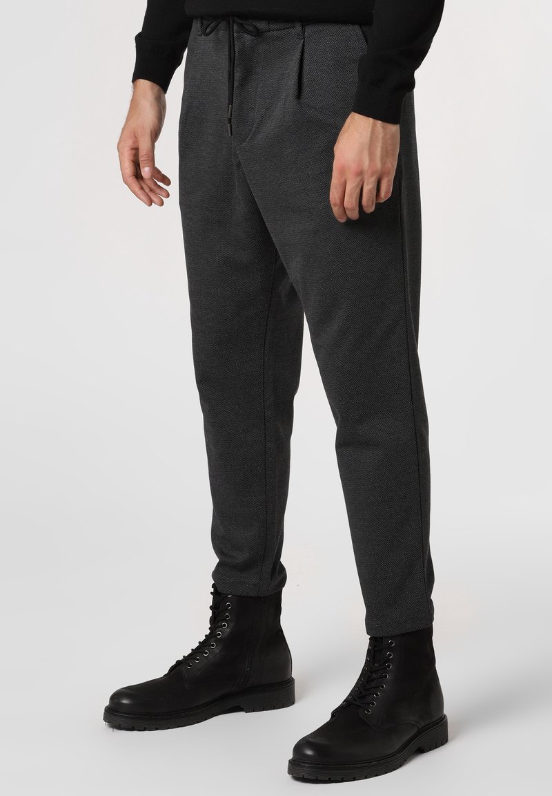 Tmavě šedé jogger kalhoty vyrobené z texturované tkaniny, s elastickým pasem, tkaničkou a postranními švy. V kombinaci s černými šněrovacími botami.