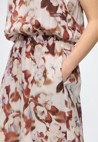 Bras plié avec la main dans la poche d'une robe à motif floral comprenant des couleurs rouge, marron, blanc et bleu clair et une taille froncée.