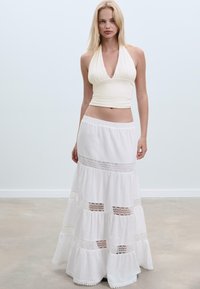 Bílý halter top spárovaný s dlouhou bílou sukní s vrstveným designem, ozdobenou krajkovými akcenty a elastickým pasem. Hladká látka s volným střihem.