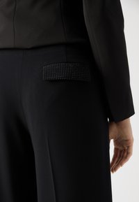 Black tailored pants featuring a textured pocket with a black studded design, smooth fabric, and a fitted shape around the waist and hips.