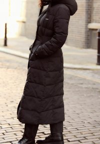 Schwarzer Steppmantel mit langem Schnitt, horizontale Steppmusterung und Kapuze. Verfügt über Seitentaschen und wird mit einem Reißverschluss geschlossen.