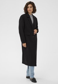 Femme aux cheveux courts et bouclés portant un long manteau noir par-dessus un pull gris, un jean bleu clair et des mocassins noirs, debout devant un fond uni.