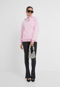 Un sweat à capuche rose clair avec poche avant et cordons de serrage, associé à un pantalon noir évasé et un sac à main texturé argenté. Des lunettes de soleil carrées noires sont portées.