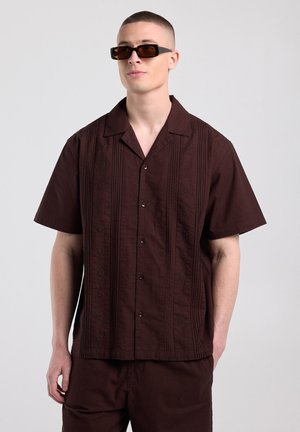 Homme aux cheveux courts portant une chemise marron foncé à manches courtes boutonnée et un pantalon assorti, accessoirisé avec des lunettes de soleil rectangulaires foncées.