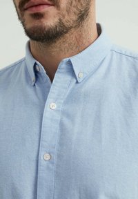 Chemise bleu clair à boutons avec un col italien, ornée de boutons blancs et d'un tissu texturé. Vue rapprochée du col et du haut de la poitrine.