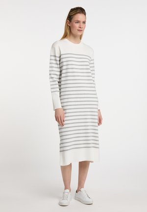 Vrouw draagt een witte midi-jurk met horizontale grijze strepen en witte sneakers, staand tegen een effen witte achtergrond.