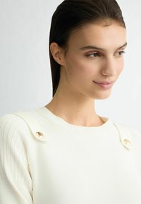 Pull blanc en tricot avec texture côtelée et accents de boutons sur l'épaule. Design col rond, tissu doux et subtile brillance.