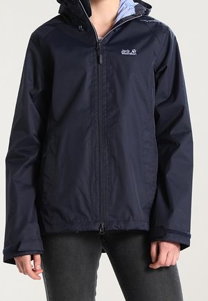 Veste d'extérieur bleu marine à capuche avec fermeture éclair à l'avant et logo sur la poitrine, portée sur un pantalon gris foncé, montrée sur la partie supérieure du corps.