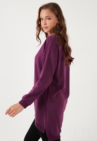 Felpa viola oversize a maniche lunghe. Tessuto morbido, scollatura rotonda, fessure laterali per facilitare i movimenti e polsini a coste. Design minimalista.