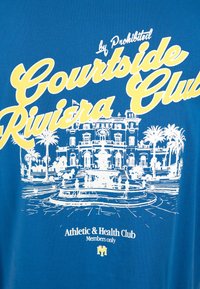 Texte jaune et blanc « Courtside Riviera Club » sur un croquis blanc d'une villa avec des palmiers et une fontaine sur un tissu bleu.
