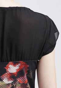 Zwarte transparante blouse met korte pofmouwen en bloemenprint rond de taille. Heeft een geraffineerde textuur met een gladde afwerking.