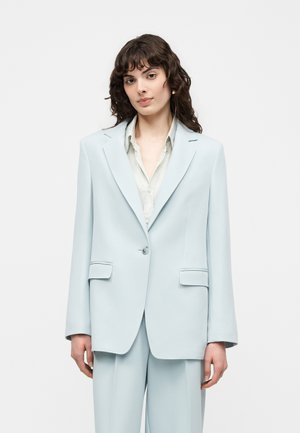 Naisel lokkis tume juustega, kes kannab helesinist suurte lõigetega bleiserit ja sobivaid pükse koos valge nööbitava särgiga.