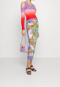 Bunter gerippter Pullover, der von Lila zu Rot übergeht, kombiniert mit gemusterten pastellfarbenen Hosen mit goldenen Akzenten. Eine kleine lavendelfarbene Handtasche rundet den Look ab.