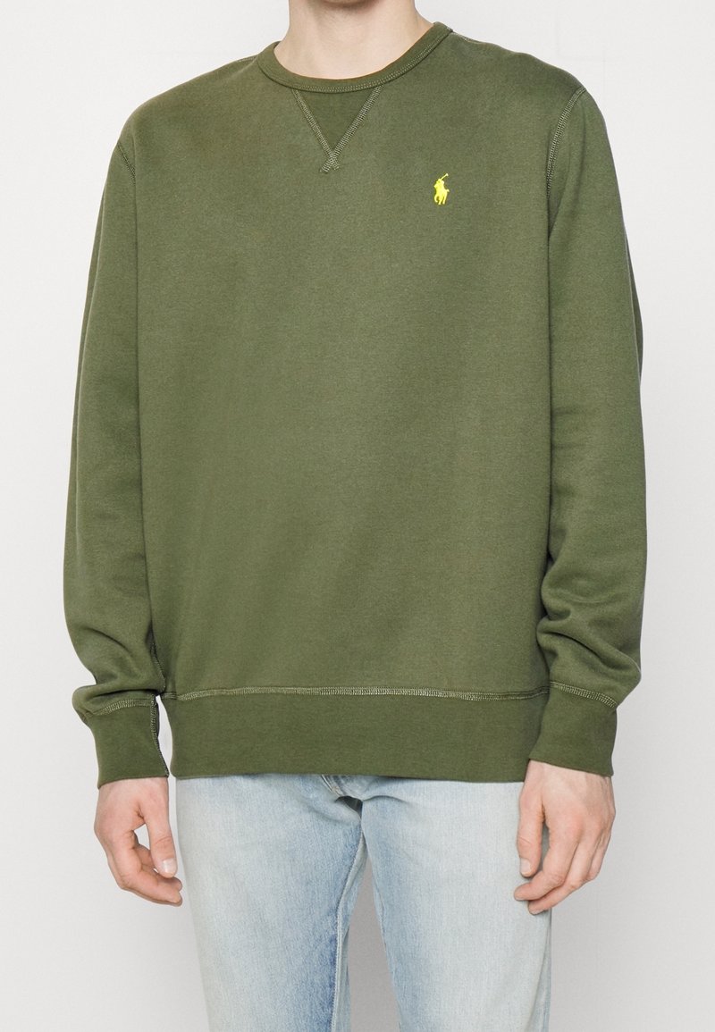 Personne portant un sweat-shirt vert olive à manches longues avec un petit logo jaune brodé sur la poitrine gauche et un jean bleu clair.
