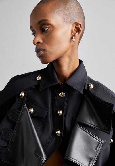 Black jacket featuring gold dome buttons, large pockets, and leather accents. Structured design with a collared neckline and a smooth texture.