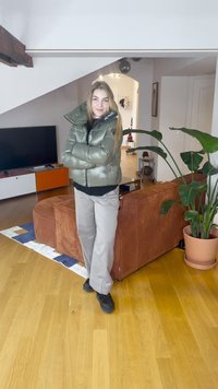 Doudoune vert métallisé, coupe cintrée, avec col montant. Pantalon gris clair et chaussures noires. Intérieur avec des plantes.