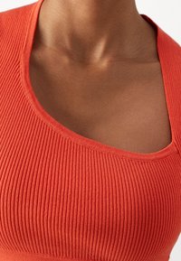 Zbliżenie osoby noszącej pomarańczowy sweter z prążkowanego dzianiny z dekoltem w serek, ukazujące górną część klatki piersiowej i obszar obojczyków.