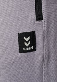 Material sportiv de culoare gri, cu o suprafață texturată, având o aplicație pătrată neagră etichetată "hummel" și două chevrone albe deasupra logo-ului.