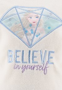 La parte superior de forro polar presenta un diseño en forma de diamante con una superposición transparente, exhibiendo una ilustración de un personaje y el texto "CREE en ti mismo" bordado en azul y púrpura.