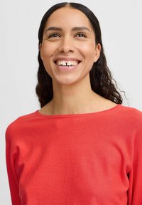 Pull rouge à col rond, fabriqué en tissu doux. Le modèle a de longs cheveux ondulés et un large sourire authentique, sur fond clair.