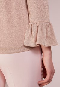 Haut rose clair avec une texture douce et brillante et des manches à volants. Associé à un pantalon rose pâle, présentant un tissu lisse et uniforme.
