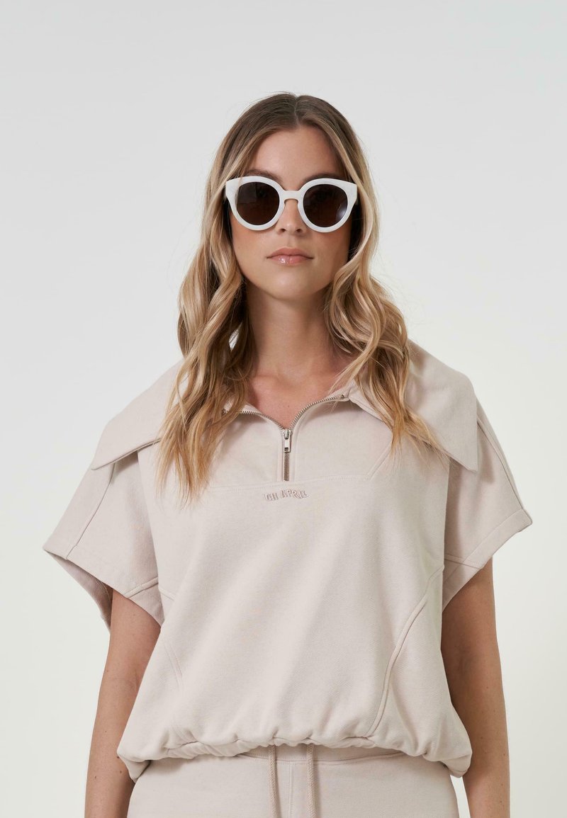 Haut zippé beige clair en tissu doux, avec des manches courtes, une taille froncée et un discret logo. Associé à des lunettes de soleil blanches en forme de yeux de chat.