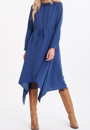 Vrouw die een blauwe midi-jurk met lange mouwen en een puntige zoom draagt, samen met beige kniehoge laarzen, staand tegen een witte achtergrond.