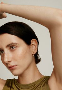 Pendiente de aro de oro con una pequeña piedra transparente. La modelo tiene el cabello corto y oscuro, lleva una blusa sin mangas verde sobre un fondo neutro.
