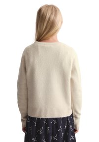 Maglione color crema con finitura testurizzata, scollo rotondo e polsini e orlo a coste, indossato sopra un vestito blu navy con motivi a fiocco.