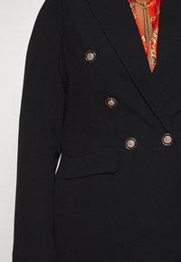 Blazer negro de doble botonadura con botones dorados ornamentados, llevado sobre una camisa roja estampada y cadenas doradas superpuestas.