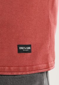 T-shirt rossa in cotone con texture a coste, con un'etichetta del marchio nera "ONLY & SONS" con testo bianco sull'orlo inferiore.