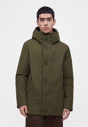 Jeune homme portant un manteau d'hiver vert à capuche avec de gros boutons, debout devant un fond blanc uni.