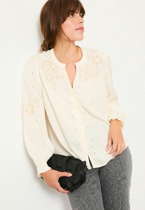 Cream, long-sleeve blouse with floral embroidery, button-up front, gathered cuffs, worn with patterned gray pants and a black crossbody bag.