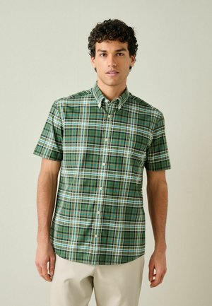 Junger Mann, der ein kariertes Hemd mit kurzen Ärmeln in Grün, Blau und Weiß sowie beige Hosen trägt, vor einem schlichten Hintergrund stehend.