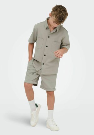 Dreng iført lysegrå kortærmet skjorte med knapper og forlommer samt tilhørende shorts, hvide sokker og hvide sneakers, stående på hvid baggrund.