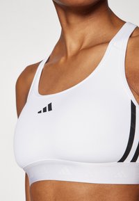 Witte sportbeha met een gladde textuur, voorzien van het zwarte adidas-logo, verticale zwarte strepen langs de zijkanten en een witte elastische band aan de onderkant.
