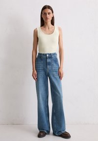 Vrouw staat tegen een witte muur met een mouwloos lichtbeige top, hoogwaardige wijdvallende blauwe jeans en donkere bruine schoenen.