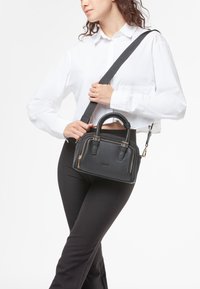 Borsa a mano in pelle nera con superficie texturizzata, doppie maniglie superiori, tracolla rimovibile e zip dorate. Design compatto e rettangolare.