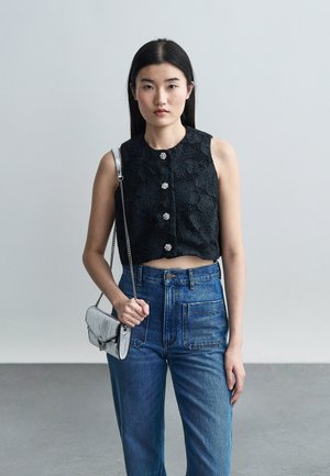 Joven con blusa sin mangas negra de encaje con botones decorativos, jeans altos de color azul y llevando un pequeño bolso plateado.