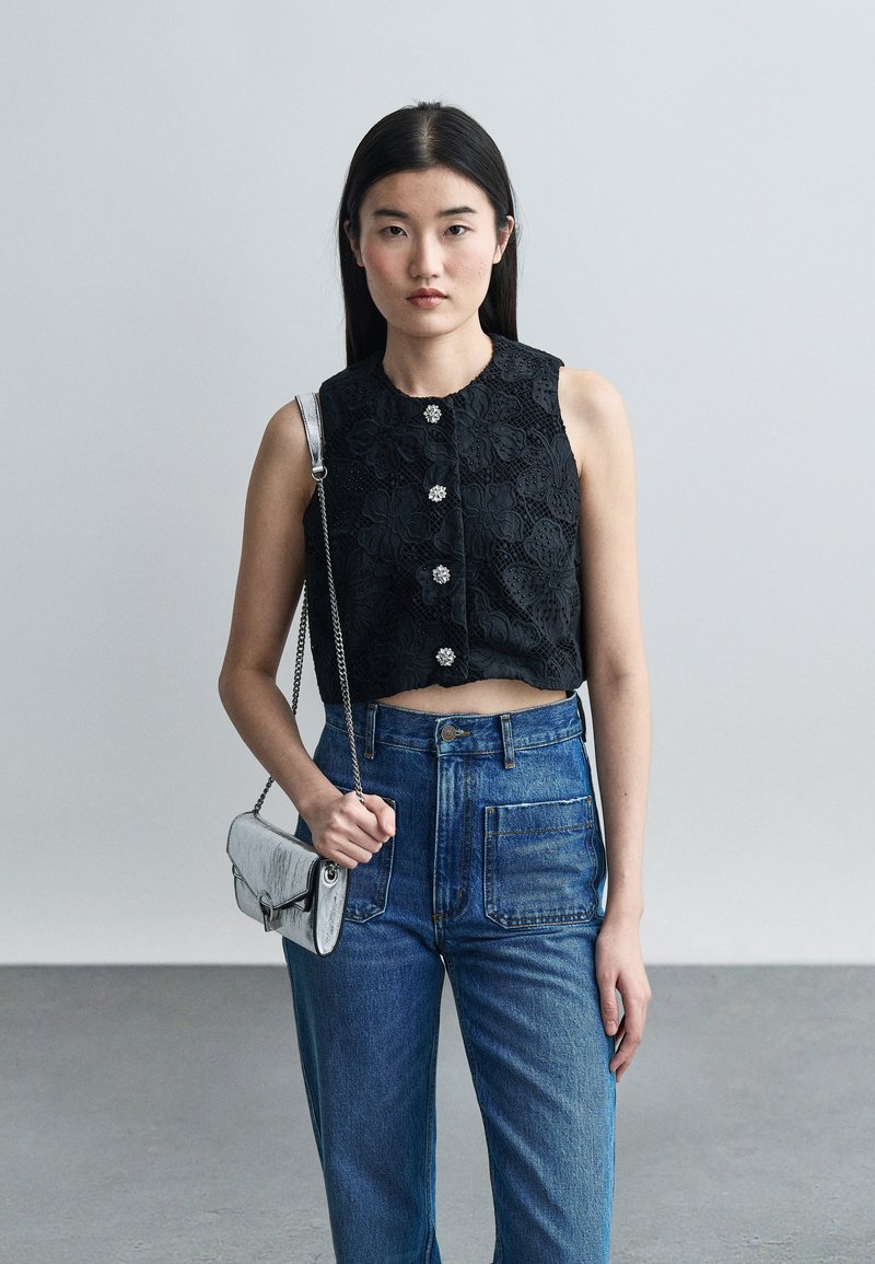 Νέα γυναίκα που φοράει μαύρο δαντελένιο τοπ χωρίς μανίκια με διακοσμητικά κουμπιά, ψηλόμεσα μπλε τζιν και κρατά μια μικρή ασημένια τσάντα.