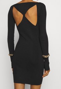 Czarna, prążkowana sukienka z długimi rękawami, z odkrytymi plecami i unikalnym wzorem wycięć. Złoty łańcuszek jako ozdoba na mankietach rękawów.