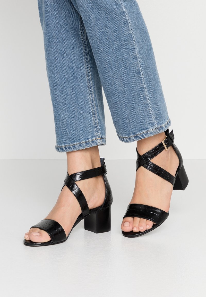 Pieds portant des sandales à talons carrés noires à motif croco avec des brides à la cheville, assorties à un jean en denim bleu.