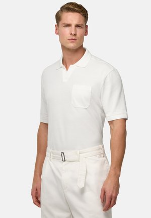Weißes Poloshirt mit kurzen Ärmeln, aus strukturiertem Stoff und mit einer Brusttasche. Getragen mit hellen Hosen und einem Gürtel.