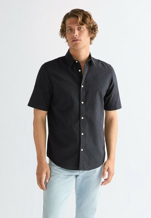 Camisa negra de manga corta, con textura de rayas verticales, cuello abotonado y botones de color claro, combinada con denim azul claro.