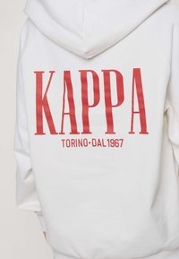 Valge kapuutsiga dressipluus, millel on seljal suur punane tekst "KAPPA" ja väiksem tekst "TORINO DAL 1967", mida kannab seljaga vaataja poole olev inimene.